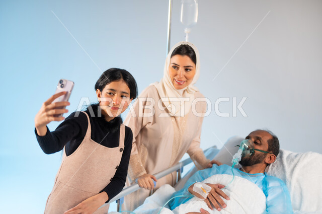 التقاط صور سيلفي تذكارية عبر الهاتف المحمول، استخدام الأجهزة والتقنيات الحديثة المتطورة، إجراء مكالمة فيديو عن طريق الجوال، بورتريه مقرب لرجل عربي خليجي سعودي يرتدي يونيفورم العمليات مستلقي على السرير يقفون بجانبه عائلته, مفهوم الطب والرعاية الصحية، خلفية زرقاء