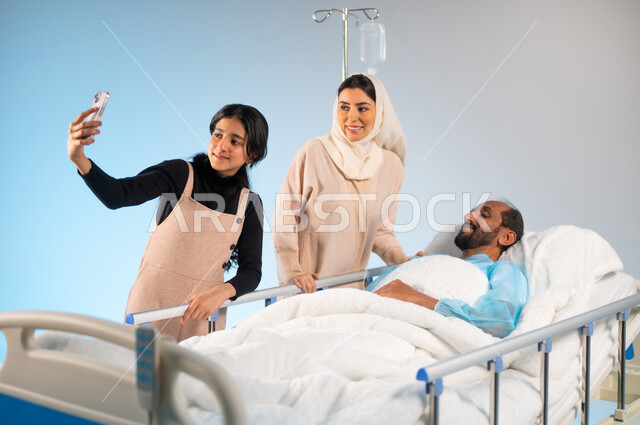 ايماءات الفرح والسرور، التقاط صورة تذكارية والنظر الى الهاتف المحمول ،  بورتريه لرجل عربي خليجي سعودي يرتدي يونيفورم العمليات مستلقي على السرير يقفون بجانبه زوجته وابنته, مفهوم الطب والرعاية الصحية، تلقي العلاج والعناية في المستشفى، خلفية زرقاء