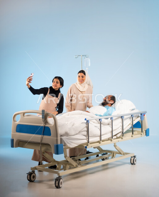 التقاط صورة تذكارية والنظر الى الهاتف المحمول ،  بورتريه لرجل عربي خليجي سعودي يرتدي يونيفورم العمليات مستلقي على السرير يقفون بجانبه زوجته وابنته, ايماءات الفرح والسرور، مفهوم الطب والرعاية الصحية، تلقي العلاج والعناية في المستشفى، خلفية زرقاء