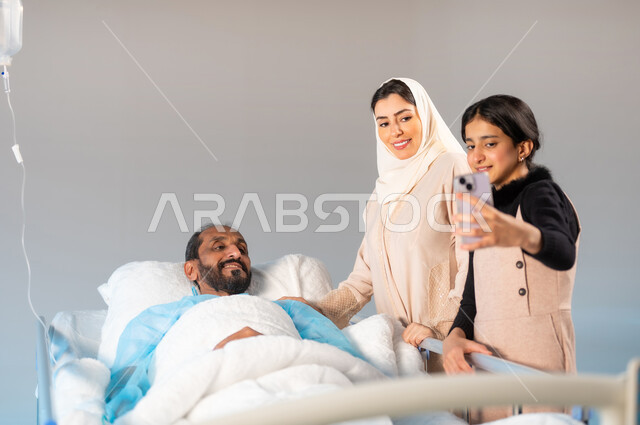 النظر الى الهاتف المحمول والتقاط صورة تذكارية، مفهوم الطب والرعاية الصحية، تلقي العلاج والعناية في المستشفى، بورتريه مقرب لرجل عربي خليجي سعودي يرتدي يونيفورم العمليات مستلقي على السرير يقفون بجانبه زوجته وابنته, ايماءات الفرح والسرور، خلفية زرقاء