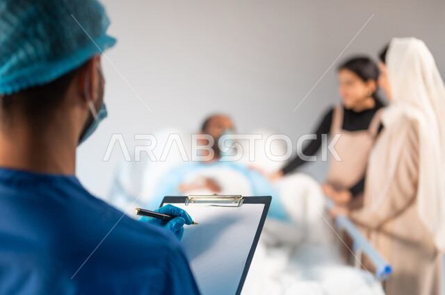 الاهتمام بالمرضى والتشخيص العلاجي لهم, تدوين المعلومات والملاحظات الطبية, صورة ضبابية مقربة لممرض عربي خليجي سعودي يرتدي يونيفورم العمل والكمامة, تلقي العلاج والعناية في المستشفى, تقديم الاستشارات والخدمات العلاجية، مفهوم الطب والرعاية الصحية، خلفية زرقاء