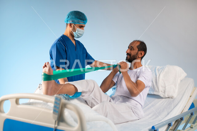 العمل في المراكز الطبية والمستشفيات السعودية، تلقي العلاج والعناية بالمستشفى، مفهوم الطب والرعاية الصحية، بورتريه لممرض عربي خليجي سعودي يرتدي بونيفورم العمل والكمامة يقوم بتقديم العلاج الفيزيائي للمريض، تقديم الاستشارات والخدمات العلاجية للمرضى، خلفية زرقاء