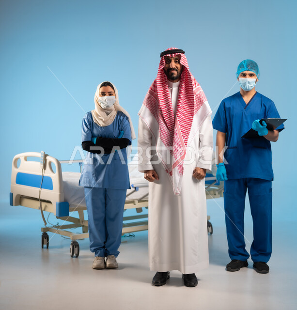 مهن ووظائف سعودية, الوقوف باستقامة والنظر إلى الكاميرا, جودة الخدمات الطبية في السعودية, بورتريه لرجل عربي خليجي سعودي برفقة ممرض وممرضة يرتديان يونيفورم العمل في إحدى المستشفيات, متابعة الحالات المرضية والتشخيص العلاجي للمرضى, صورة الجسم بالكامل, خلفية زرقاء