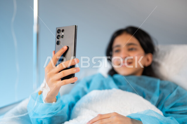 التقاط صور سيلفي تذكارية عبر الهاتف المحمول, إجراء مكالمة فيديو عبر الجوال, بورتريه مقرب لفتاة مريضة عربية خليجية سعودية مبتسمة ترتدي يونيفورم العمليات تستلقي على السرير, استخدام الأجهزة والتقنيات الحديثة والمتطورة, خلفية زرقاء