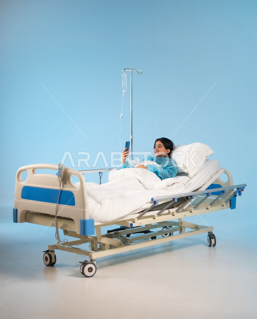 إجراء مكالمة فيديو عبر الجوال, بورتريه لفتاة مريضة عربية خليجية سعودية مبتسمة ترتدي يونيفورم العمليات تستلقي على السرير, التقاط صور سيلفي تذكارية عبر الهاتف المحمول, استخدام الأجهزة والتقنيات الحديثة والمتطورة, خلفية زرقاء