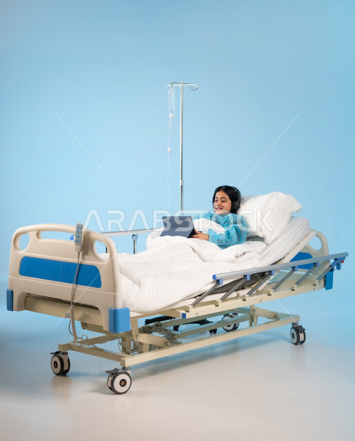 الترفيه والتسلية عبر التطبيقات والبرامج الالكترونية, استخدام الأجهزة والتقنيات الحديثة والمتطورة, بورتريه لفتاة مريضة عربية خليجية سعودية مبتسمة ترتدي الهيدفون يونيورم العمليات تمسك بيدها جهاز التابليت, مستشفيات خاصة بالأطفال في المملكة, خلفية زرقاء