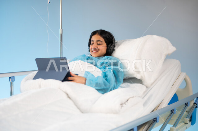 متابعة الأفلام والبرامج الترفيهية عبر التطبيقات الالكترونية, بورتريه لفتاة مريضة عربية خليجية سعودية ترتدي الهيدفون ويونيفورم العمليات مستقلية على السرير تحمل بيدها جهاز التابليت, استخدام الأجهزة والتقنيات الحديثة والمتطورة, مستشفيات خاصة للأطفال في المملكة, خلفية زرقاء