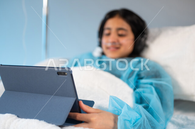 التسلية والترفيه عبر التطبيقات والبرامج الالكترونية, استخدام الأجهزة والتقنيات الحديثة والمتطورة, بورتريه ضبابي مقرب لفتاة مريضة عربية خليجية سعودية مبتسمة ترتدي الهيدفون ويونيورم العمليات تمسك بيدها جهاز التابليت, مستشفيات خاصة بالأطفال في المملكة, خلفية زرقاء