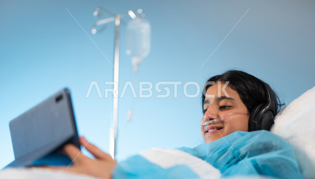 استخدام الأجهزة والتقنيات الحديثة والمتطورة, بورتريه مقرب لفتاة مريضة عربية خليجية سعودية مبتسمة  ترتدي الهيدفون ويونيورم العمليات تمسك بيدها جهاز التابليت, مستشفيات خاصة بالأطفال في المملكة, التسلية والترفيه عبر التطبيقات والبرامج الالكترونية, خلفية زرقاء