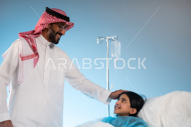 نظرات مليئة بالعطف والحنان والاهتمام, تلقي العلاج والعناية في المستشفى, مفهوم العناية الطبية والخوف على صحة الأبناء, بورتريه لرجل عربي خليجي سعودي يرتدي الثوب التقليدي والشماغ يقف بجانب ابنته المستلقية على السرير, خلفية زرقاء