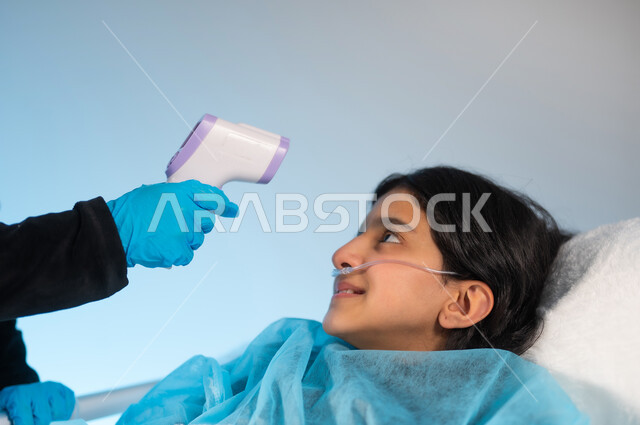 اتباع الاجراءات الاحترازية للوقاية من فايروس كورونا, بورتريه مقرب ليد ممرضة تقوم بفحص درجة حرارة الجسم لفتاة عربية خليجية سعودية ترتدي بونيفورم العمليات مستلقية على سرير في المستشفى, مفهوم الطب والرعاية الصحية, خلفية زرقاء