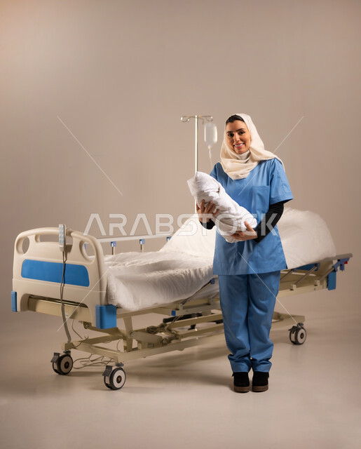 الوقوف باستقامة والنظر إلى الكاميرا, مهن ووظائف نسائية طبية, الاعتناء بالأطفال الرضع حديثي الولادة, بورتريه لممرضة عربية خليجية سعودية ترتدي يونيفورم العمل تحمل بيدها طفل رضيع, مفهوم الطب والرعاية الصحية, صورة الجسم بالطول الكامل, خلفية بيج