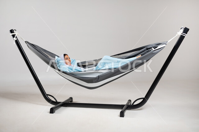 ايماءات تدل على الراحة والسرور، الاسترخاء والتأمل في اوقات الفراغ، بورتريه لشابة عربية خليجية سعودية ترتدي لباس الكاجوال الخاص بالرحلات الصيفية مستلقية على أرجوحة قماشية تضع يديها خلف رأسها و تنظر الى الأعلى، متعة قضاء الوقت مع الذات، خلفية رمادية