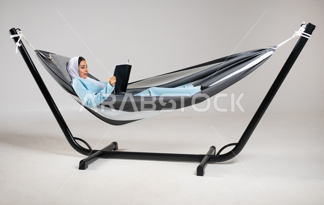 قضاء الوقت بعيداً عن التكنولوجيا، متعة قضاء الوقت مع الذات، بورتريه لشابة عربية خليجية سعودية ترتدي لباس الكاجوال الخاص بالرحلات الصيفية مستلقية على أرجوحة قماشية تمسك بيدها مجلة ثقافية، استغلال وقت الاستراحة بالمطالعة، ممارسة هواية قراءة المجلات، خلفية رمادية