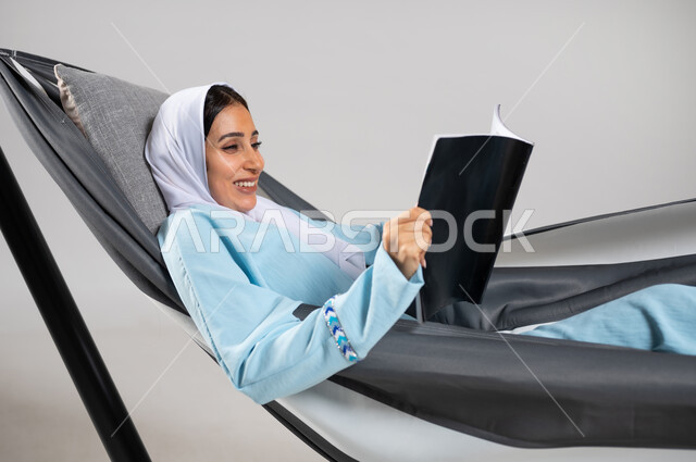 استغلال وقت الاستراحة بالمطالعة، ممارسة هواية قراءة المجلات، بورتريه مقرب لشابة عربية خليجية سعودية ترتدي لباس الكاجوال الخاص بالرحلات الصيفية مستلقية على أرجوحة قماشية تمسك مجلة ثقافية، قضاء الوقت بعيداً عن التكنولوجيا، متعة قضاء الوقت مع الذات، خلفية رمادية