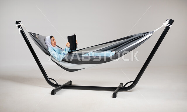 Practicing the hobby of reading magazines, using the break time to read, a portrait of a Saudi Gulf Arab young woman wearing casual clothes for summer trips, lying on a cloth hammock holding a cultural magazine in her hand, spending time away from technology, the pleasure of spending time with oneself, gray background