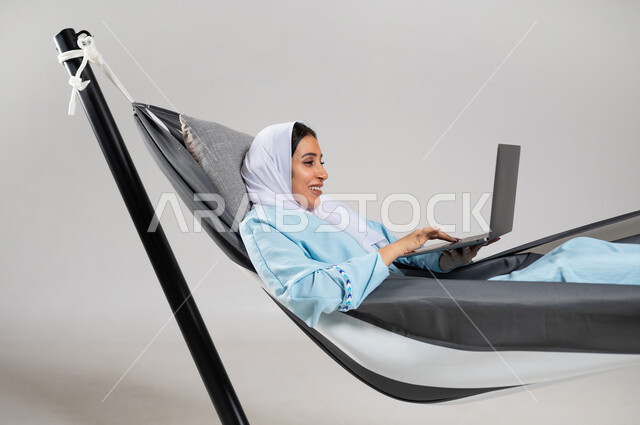 تصفح الانترنت و وسائل التواصل الاجتماعي، إنجاز المهام عن بعد، استخدام التقنية والتكنولوجيا الحديثة، بورتريه مقرب لشابة عربية خليجية سعودية ترتدي لباس الكاجوال الخاص بالرحلات الصيفية مستلقية على أرجوحة قماشية تقوم بالعمل على جهاز الحاسب المحمول، خلفية رمادية