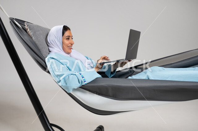 استخدام التقنية والتكنولوجيا الحديثة، تصفح الانترنت و وسائل التواصل الاجتماعي، إنجاز المهام عن بعد، بورتريه مقرب لشابة عربية خليجية سعودية ترتدي لباس الكاجوال الخاص بالرحلات الصيفية مستلقية على أرجوحة قماشية تقوم بالعمل على جهاز الحاسب المحمول، خلفية رمادية