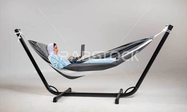 تصفح الانترنت و وسائل التواصل الاجتماعي باستخدام التقنيات الحديثة والمتطورة، إنجاز المهام عن بعد، بورتريه لشابة عربية خليجية سعودية ترتدي لباس الكاجوال الخاص بالرحلات الصيفية مستلقية على أرجوحة قماشية تقوم بالعمل على جهاز الحاسب المحمول، خلفية رمادية