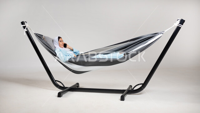 تبادل الأحاديث والأخبار الجيدة، التواصل مع الأهل والأصدقاء، الاتصال الهاتفي عبر الجوال، بورتريه لشابة عربية خليجية سعودية ترتدي لباس الكاجوال الخاص بالرحلات الصيفية مستلقية على أرجوحة قماشية معلقة تقوم بإجراء مكالمة هاتفية عبر الهاتف المحمول، خلفية رمادية