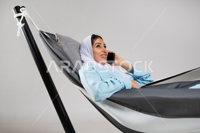 التواصل مع الأهل والأصدقاء، تبادل الأحاديث والأخبار الجيدة، الاتصال الهاتفي عبر الجوال، بورتريه مقرب لشابة عربية خليجية سعودية ترتدي لباس الكاجوال الخاص بالرحلات الصيفية مستلقية على أرجوحة قماشية معلقة تقوم بإجراء مكالمة هاتفية عبر الهاتف المحمول، خلفية رمادية