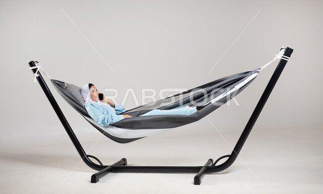 Mobile phone calls, communication with family and friends, exchanging conversations and good news, portrait of a Saudi Gulf Arab woman wearing casual clothes for summer trips, lying on a hanging cloth hammock making a phone call via mobile phone, gray background