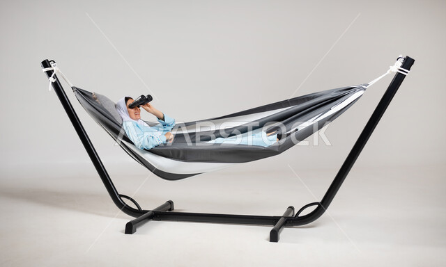 Spending time relaxing and calm, a portrait of a Saudi Gulf Arab young woman wearing casual clothes for summer trips, lying on a hanging cloth hammock, looking through binoculars with two lenses, zooming in on distant objects, gestures of entertainment, pleasure and amusement, gray background