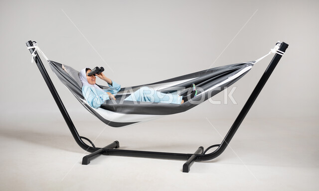 الاسترخاء والهدوء في الاستراحة، النظر من خلال المنظار المكبر بعدستين، تقريب الأشياء البعيدة، إيماءات الترفيه والمتعة والتسلية، بورتريه لشابة عربية خليجية سعودية ترتدي لباس الكاجوال الخاص بالرحلات الصيفية مستلقية على أرجوحة قماشية معلقة، خلفية رمادية