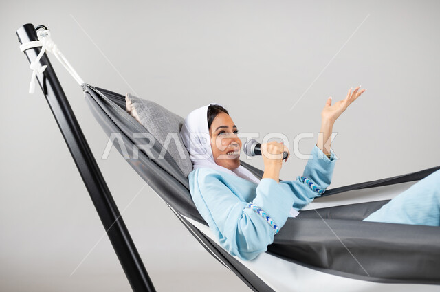 أنشطة ترفيهية مسلية، أداء المواهب الصوتية، إيماءات التفاعل والتسلية، بورتريه مقرب لشابة عربية خليجية سعودية ترتدي لباس الكاجوال الخاص بالرحلات الصيفية مستلقية على أرجوحة قماشية معلقة تقوم بالغناء باستخدام المايكروفون، متعة قضاء الوقت مع الذات، خلفية رمادية