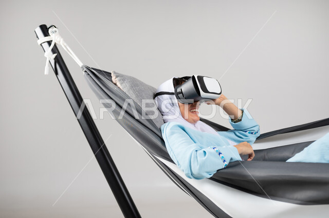 ارتداء نظارات الواقع الافتراضي ثلاثية الأبعاد، المحاولة بالتركيز والاندماج، بورتريه مقرب لشابة عربية خليجية سعودية ترتدي لباس الكاجوال الخاص بالرحلات الصيفية مستلقية على أرجوحة قماشية معلقة، إيماءات التفاعل والتسلية، متعة قضاء الوقت مع الذات، خلفية رمادية