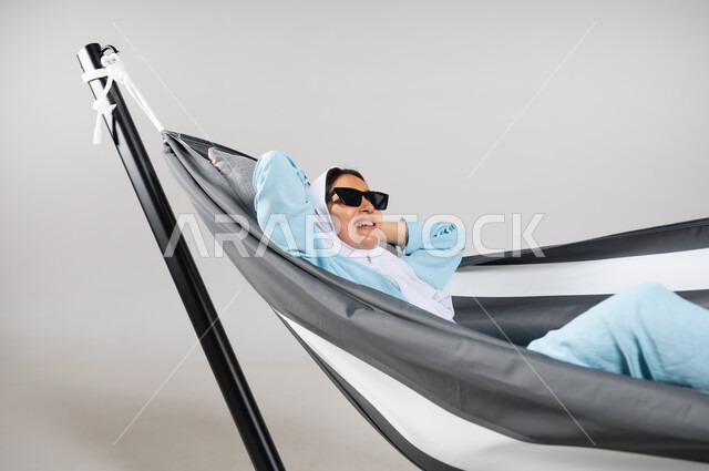 الاستجمام والراحة في أوقات الاستراحة، متعة قضاء الوقت مع الذات، بورتريه مقرب لشابة عربية خليجية سعودية ترتدي لباس الكاجوال الخاص بالرحلات الصيفية مستلقية على أرجوحة قماشية معلقة ترتدي النظارات الشمسية ذات اللون الأسود، إيماءات تدل الفرح والسرور، خلفية رمادية