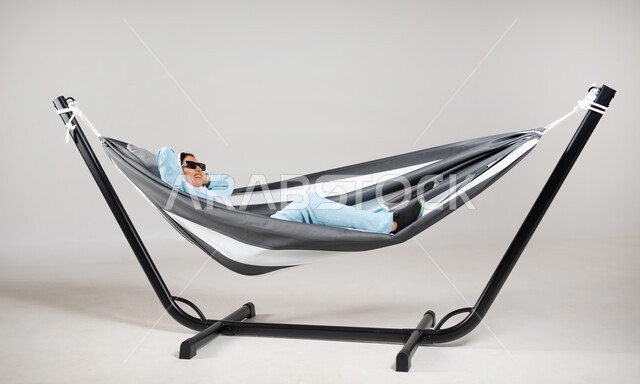 ممارسة الاسترخاء والاستجمام، بورتريه لشابة عربية خليجية سعودية ترتدي لباس الكاجوال الخاص بالرحلات الصيفية مستلقية على أرجوحة قماشية معلقة ترتدي النظارات الشمسية ذات اللون الأسود، إيماءات تدل الفرح والسرور، متعة قضاء الوقت مع الذات، خلفية رمادية