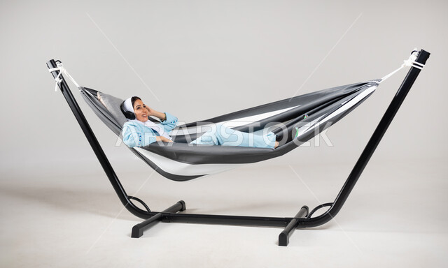 أنشطة ترفيهية مسلية، ممارسة الاستماع الى الموسيقى،عطلة صيفية ممتعة، بورتريه لشابة عربية خليجية سعودية ترتدي لباس الكاجوال الخاص بالرحلات الصيفية مستلقية على أرجوحة قماشية معلقة، إيماءات السعادة والسرور، متعة قضاء الوقت مع الذات، خلفية رمادية