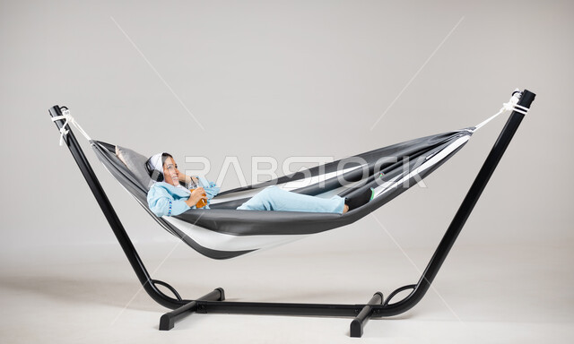 قضاء وقت ممتع بالاستماع الى الموسيقى، الاستمتاع بالمشروبات الباردة، إيماءات السعادة والفرح، أجواء صيفية سعيدة، بورتريه لشابة عربية خليجية سعودية ترتدي لباس الكاجوال الخاص بالرحلات الصيفية مستلقية على أرجوحة قماشية معلقة، متعة قضاء الوقت مع الذات، خلفية رمادية