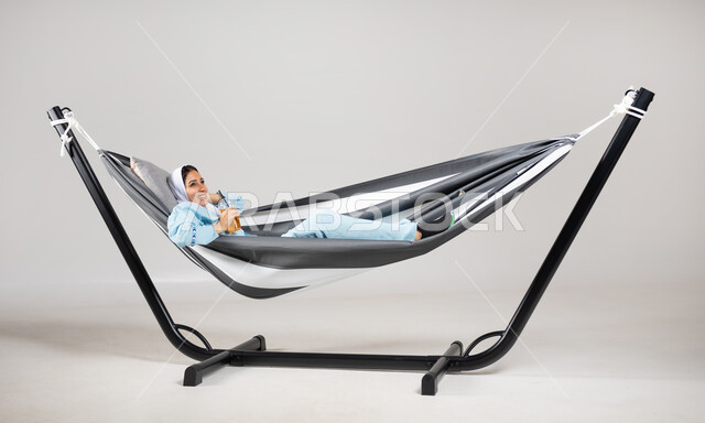 الاستمتاع بالمشروبات الباردة، إيماءات السعادة والفرح، أجواء صيفية سعيدة، بورتريه لشابة عربية خليجية سعودية ترتدي لباس الكاجوال الخاص بالرحلات الصيفية مستلقية على أرجوحة قماشية معلقة، متعة قضاء الوقت مع الذات، خلفية رمادية