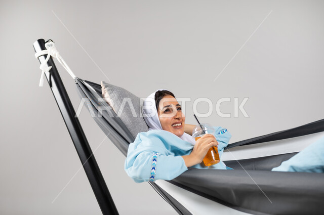 أجواء صيفية سعيدة، الاستمتاع بالمشروبات الباردة، إيماءات السعادة والفرح، متعة قضاء الوقت مع الذات، بورتريه مقرب لشابة عربية خليجية سعودية ترتدي لباس الكاجوال الخاص بالرحلات الصيفية مستلقية على أرجوحة قماشية معلقة، خلفية رمادية