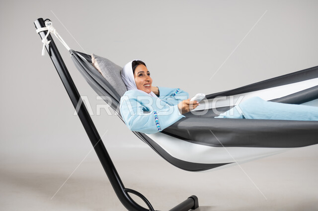 قضاء الاوقات بالتسلية والترفيه في العطلة، إيماءات السعادة والاستمتاع، متعة قضاء الوقت مع الذات، بورتريه مقرب لشابة عربية خليجية سعودية ترتدي لباس الكاجوال الخاص بالرحلات الصيفية مستلقية على أرجوحة قماشية معلقة تحمل بيدها جهاز تحكم لشيء ما، خلفية رمادية