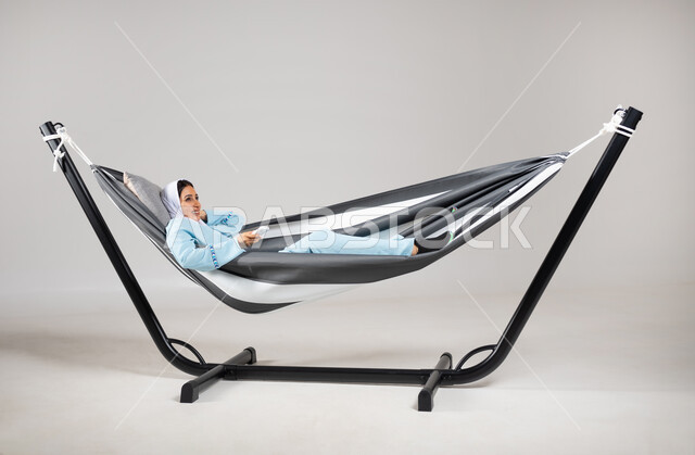 التسلية والترفيه في العطلة، إيماءات السعادة والاستمتاع، بورتريه لشابة عربية خليجية سعودية ترتدي لباس الكاجوال الخاص بالرحلات الصيفية مستلقية على أرجوحة قماشية معلقة تحمل بيدها جهاز تحكم لشيء ما، متعة قضاء الوقت مع الذات، خلفية رمادية