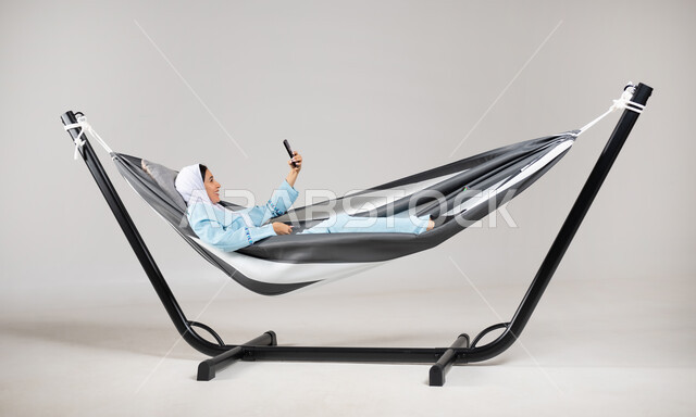 التقاط صور السيلفي من خلال الهاتف المحمول، التسلية والترفيه في العطلة، إيماءات السعادة والاستمتاع، بورتريه لشابة عربية خليجية سعودية ترتدي لباس الكاجوال الخاص بالرحلات الصيفية مستلقية على أرجوحة قماشية معلقة، متعة قضاء الوقت مع الذات، خلفية رمادية