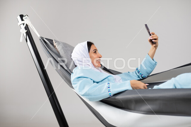 التسلية و الترفيه بالتقاط صور السيلفي من خلال الهاتف المحمول بإيماءات السعادة والاستمتاع، بورتريه مقرب لشابة عربية خليجية سعودية ترتدي لباس الكاجوال الخاص بالرحلات الصيفية مستلقية على أرجوحة قماشية معلقة، متعة قضاء الوقت مع الذات، خلفية رمادية