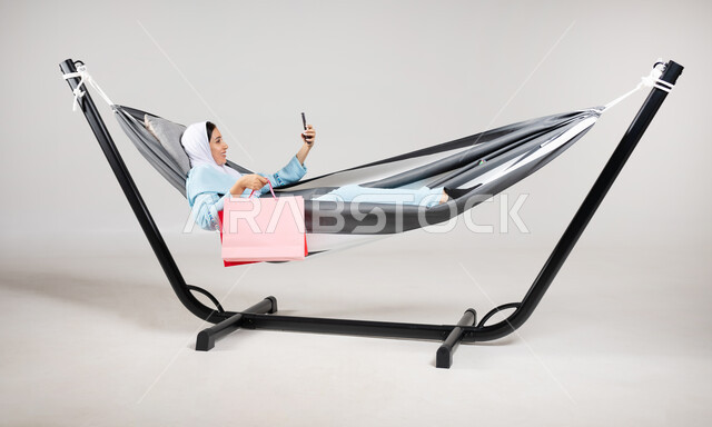 التقاط صورة سيلفي تذكارية من خلال الهاتف المحمول بإيماءات السعادة والاستمتاع، بورتريه لشابة عربية خليجية سعودية ترتدي لباس الكاجوال الخاص بالرحلات الصيفية مستلقية على أرجوحة قماشية معلقة تحمل بيدها أكياس التسوق، متعة قضاء الوقت مع الذات، خلفية رمادية