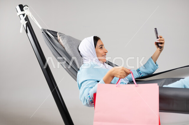 المتعة بالتقاط صورة سيلفي من خلال الهاتف المحمول بإيماءات السعادة والاستمتاع،  متعة قضاء الوقت مع الذات، بورتريه مقرب لشابة عربية خليجية سعودية ترتدي لباس الكاجوال الخاص بالرحلات الصيفية مستلقية على أرجوحة قماشية معلقة تحمل بيدها أكياس التسوق، خلفية رمادية