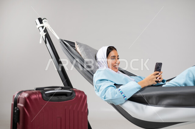 Using modern technologies to communicate, a close-up portrait of a Saudi Gulf Arab young woman wearing casual clothes for summer trips, lying on a hanging cloth hammock with a travel bag next to her, looking at the mobile phone with gestures of happiness and pleasure, the pleasure of spending time with oneself, gray background