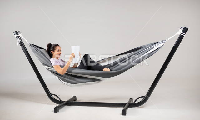 استغلال وقت الاستراحة بالفائدة، الجمع بين المتعة والفائدة، بورتريه مقرب لشابة عربية خليجية سعودية ترتدي لباس الكاجوال الخاص بالرحلات الصيفية مستلقية على أرجوحة قماشية معلقة، ايماءات السعادة و السرور، متعة قضاء الوقت مع الذات، خلفية رمادية