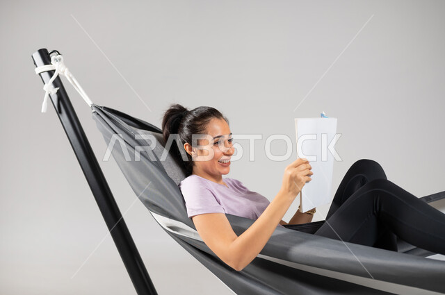 الجمع بين المتعة والفائدة، استغلال وقت الاستراحة بالفائدة، متعة قضاء الوقت مع الذات، بورتريه مقرب لشابة عربية خليجية سعودية ترتدي لباس الكاجوال الخاص بالرحلات الصيفية مستلقية على أرجوحة قماشية معلقة، ايماءات السعادة و السرور، خلفية رمادية