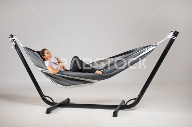 الاستمتاع بالمشروبات الصيفية الباردة، متعة قضاء الوقت مع الذات، بورتريه لشابة عربية خليجية سعودية ترتدي لباس الكاجوال الخاص بالرحلات مستلقية على ارجوحة قماشية معلقة، ايماءات السعادة و السرور، خلفية رمادية