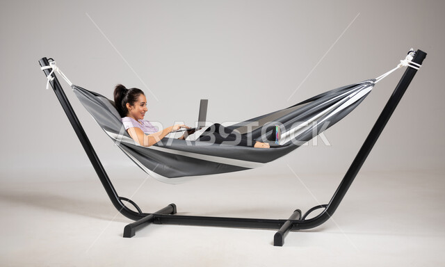 إنجاز المهام عن بعد، متعة قضاء الوقت مع الذات، بورتريه لشابة عربية خليجية سعودية ترتدي لباس الكاجوال الخاص بالرحلات الصيفية مستلقية على ارجوحة قماشية معلقة،  تصفح الانترنت و وسائل التواصل الاجتماعي باستخدام التقنيات الحديثة والمتطورة، خلفية رمادية