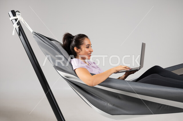 تصفح الانترنت و وسائل التواصل الاجتماعي باستخدام التقنيات الحديثة والمتطورة، إنجاز المهام عن بعد، بورتريه مقرب لشابة عربية خليجية سعودية ترتدي لباس الكاجوال الخاص بالرحلات الصيفية مستلقية على ارجوحة قماشية معلقة، متعة قضاء الوقت مع الذات، خلفية رمادية