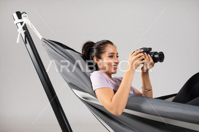 Enjoying looking at pictures in the camera, photographing the tourist trip, a close-up portrait of a Saudi Gulf Arab young woman wearing casual clothes for summer trips, lying on a hanging cloth hammock, the talent for photography and viewing souvenir pictures, a gray background