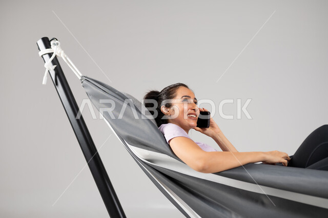 الاتصال الهاتفي عبر الهاتف المحمول، تبادل الأحاديث والأخبار الجيدة، التواصل مع الأهل والأصدقاء، بورتريه مقرب لشابة عربية خليجية سعودية مستلقية على أرجوحة قماشية معلقة ترتدي لباس الكاجوال الخاص بالرحلات الصيفية، عطلة صيفية سعيدة، خلفية رمادية